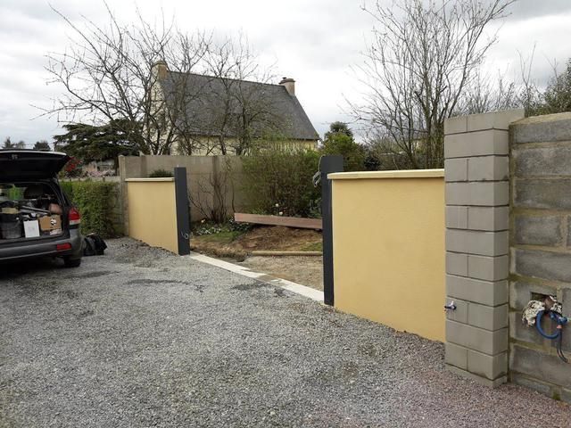 Allée de gravier longeant un mur et un portail jaunes, avec une maison et des arbres en arrière-plan.