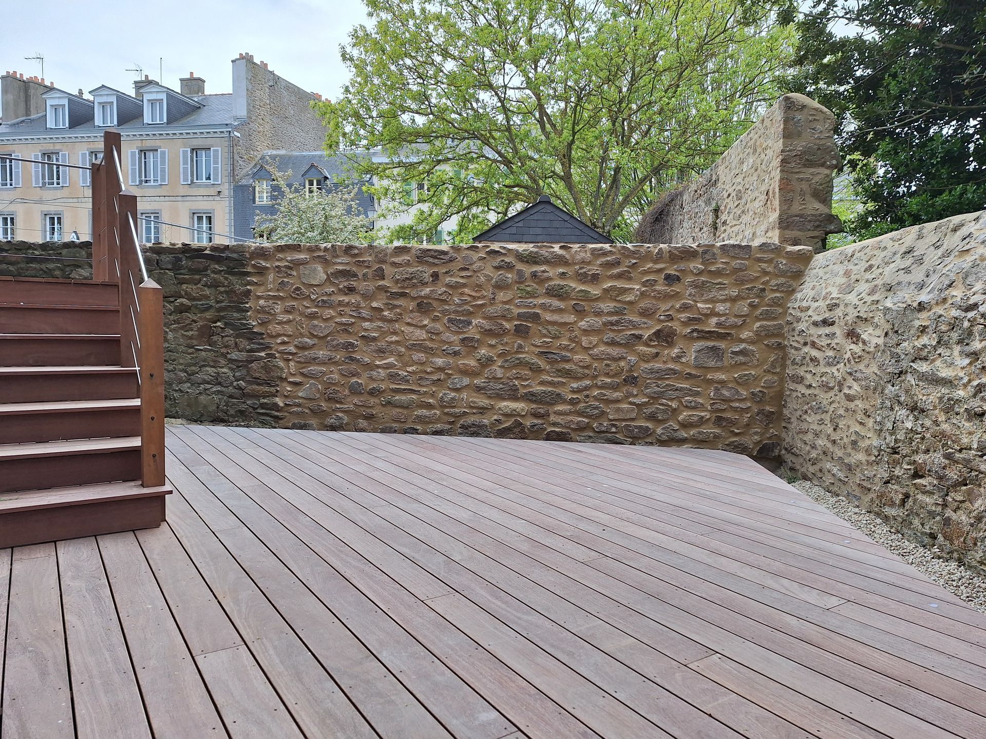 Terrasse en bois avec muret en pierre, marches et arbres donnant sur les bâtiments voisins
