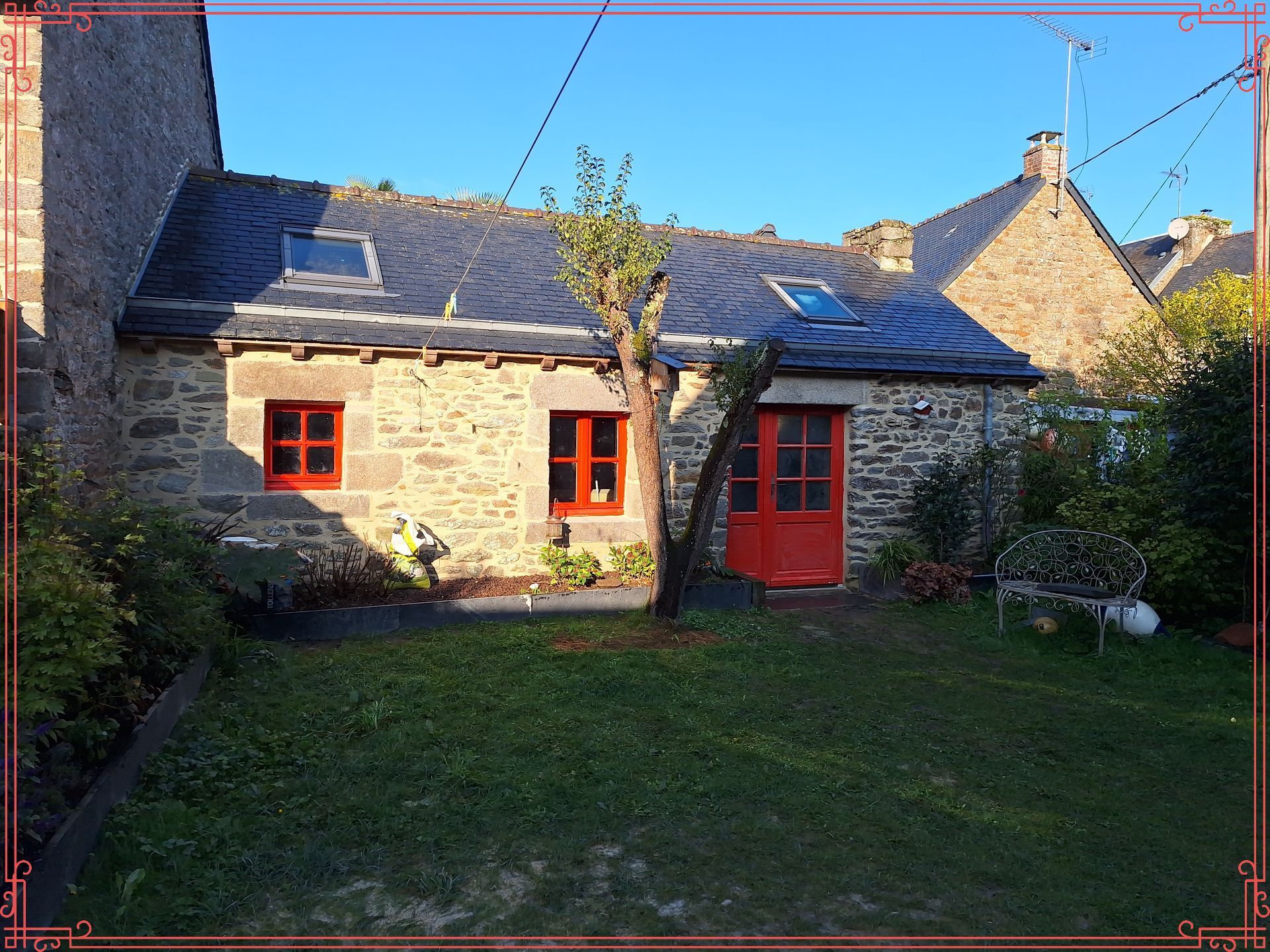 Maisonnette en pierre avec porte et fenêtres rouges, située dans un jardin ensoleillé.
