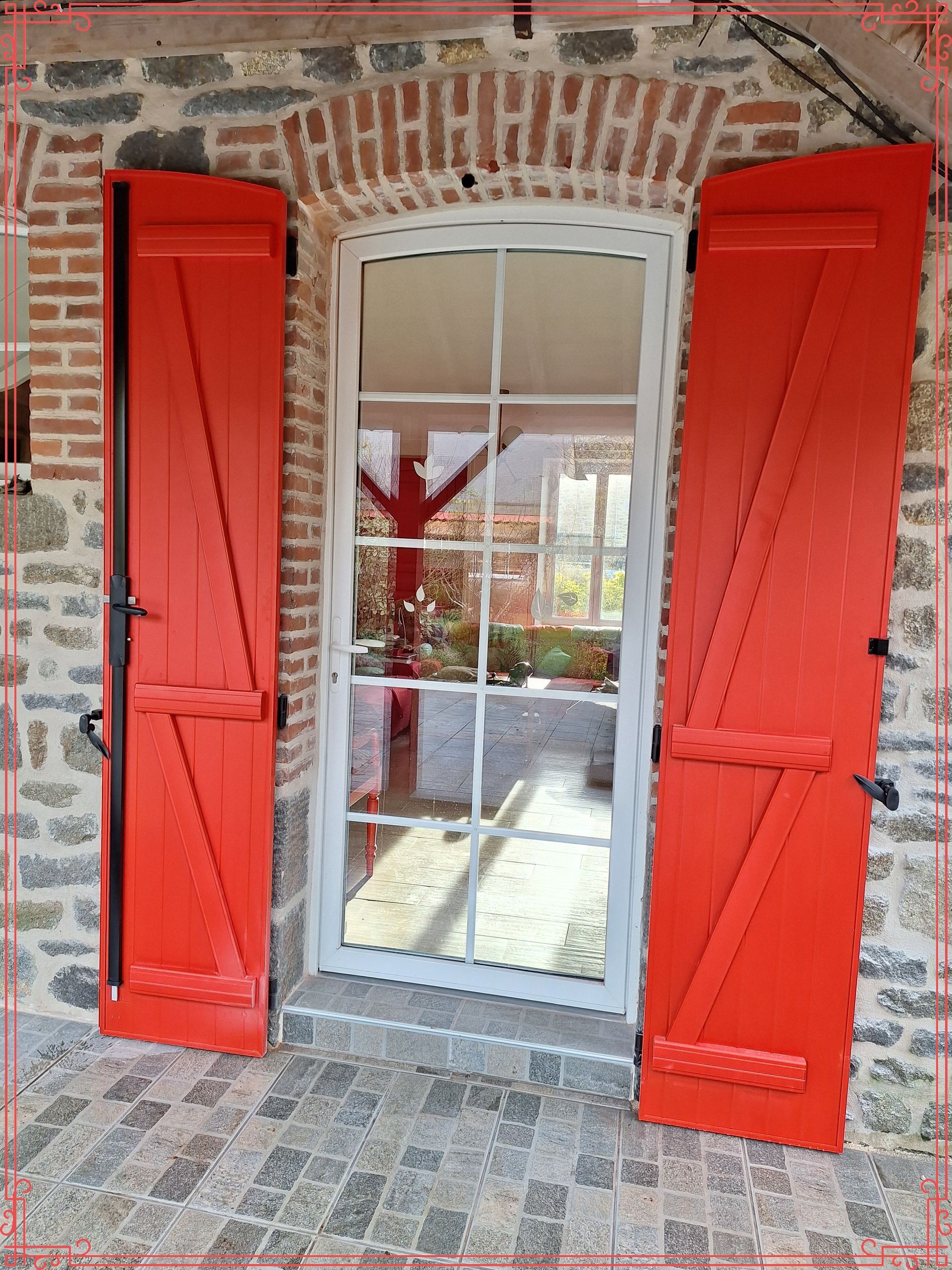 Volets rouges encadrant une porte vitrée dans un mur de briques et de pierres