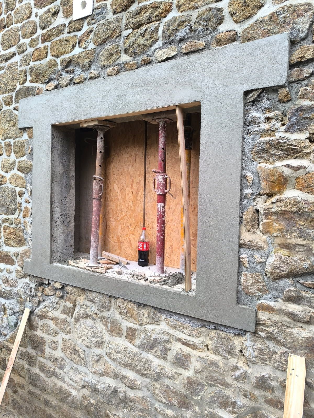 Mur en pierre percé d'une petite fenêtre encadrée de gris, laissant apparaître des planches de bois et une bouteille à l'intérieur.