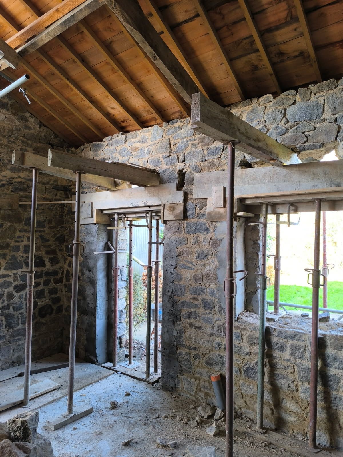 Pièce en pierre en cours de rénovation avec poutres apparentes au plafond, murs nus et ouvertures de fenêtres.