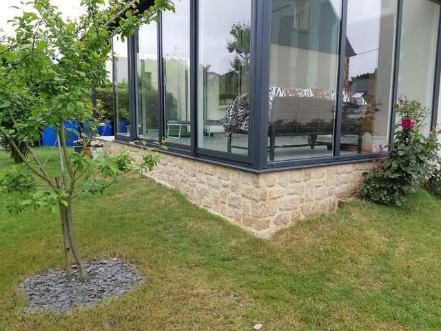 Coin d'un bâtiment vitré sur socle de pierre, pelouse et petit arbre au premier plan