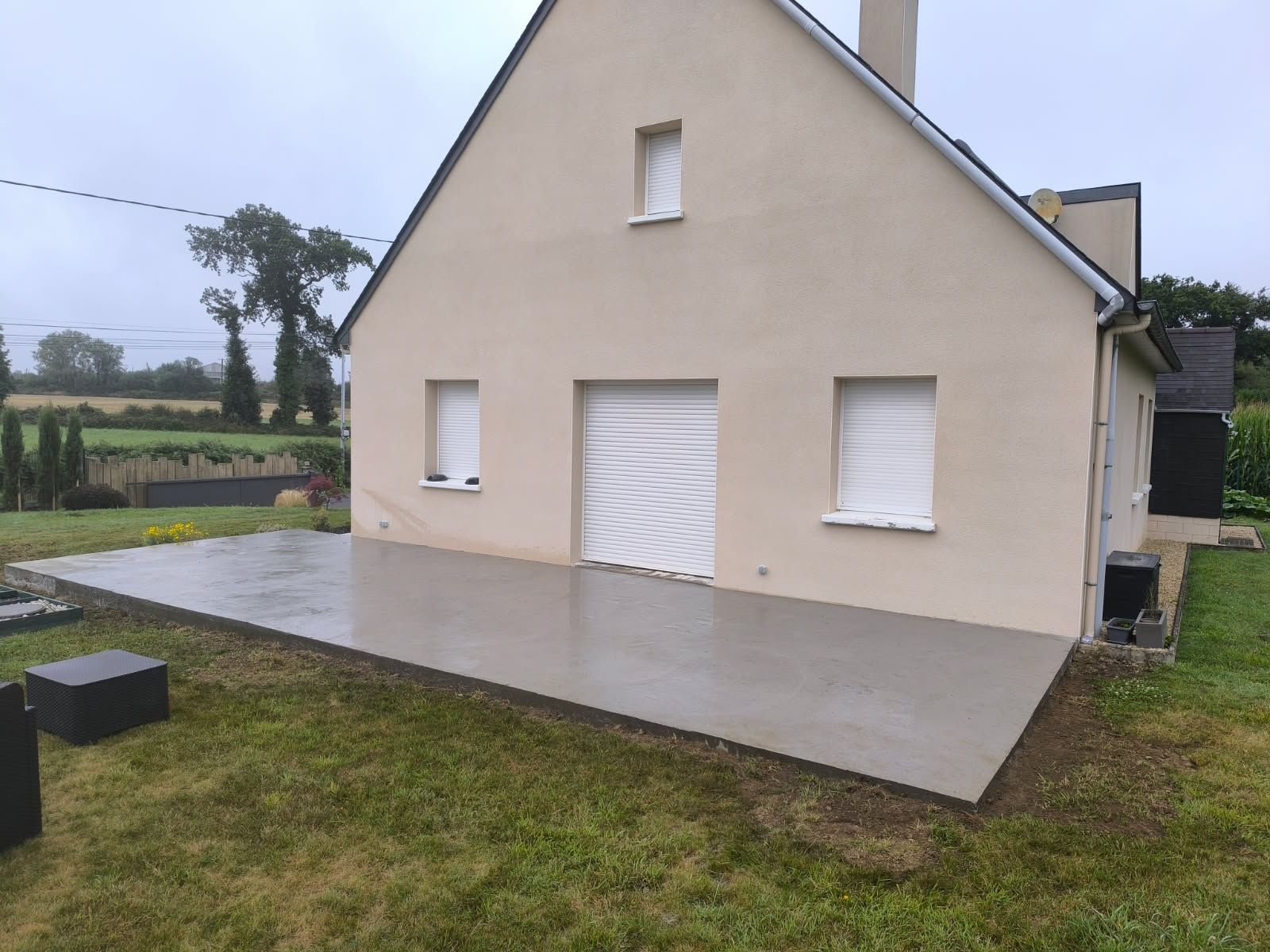 Maison beige aux volets blancs et porte de garage donnant sur une pelouse sous un ciel couvert.
