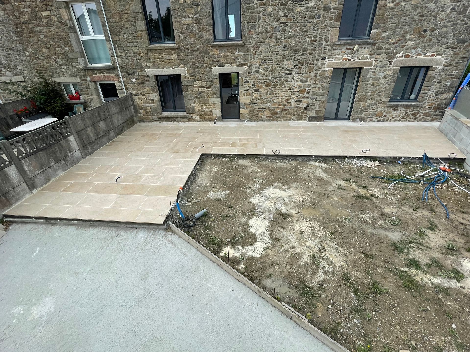 Terrasse en pierre et cour en béton jouxtant un bâtiment en briques, avec une petite parcelle de terre et un muret de soutènement.