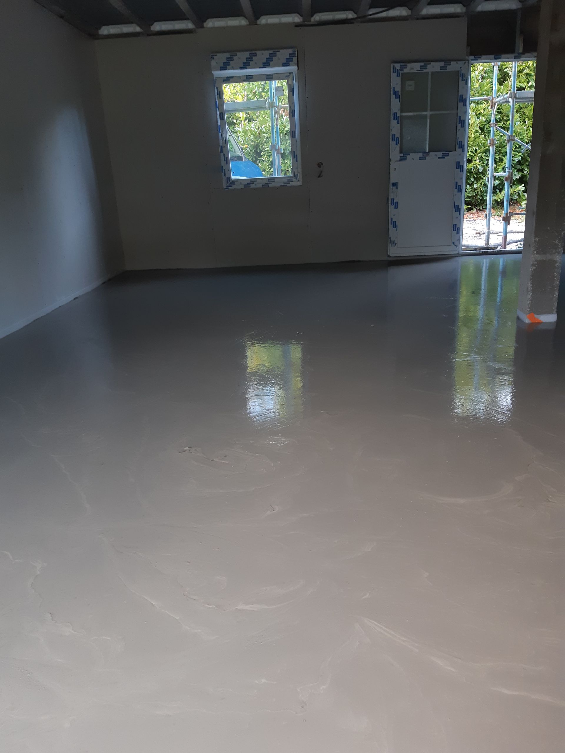 Pièce vide avec sol gris clair brillant, fenêtre et porte ouverte donnant sur la verdure.