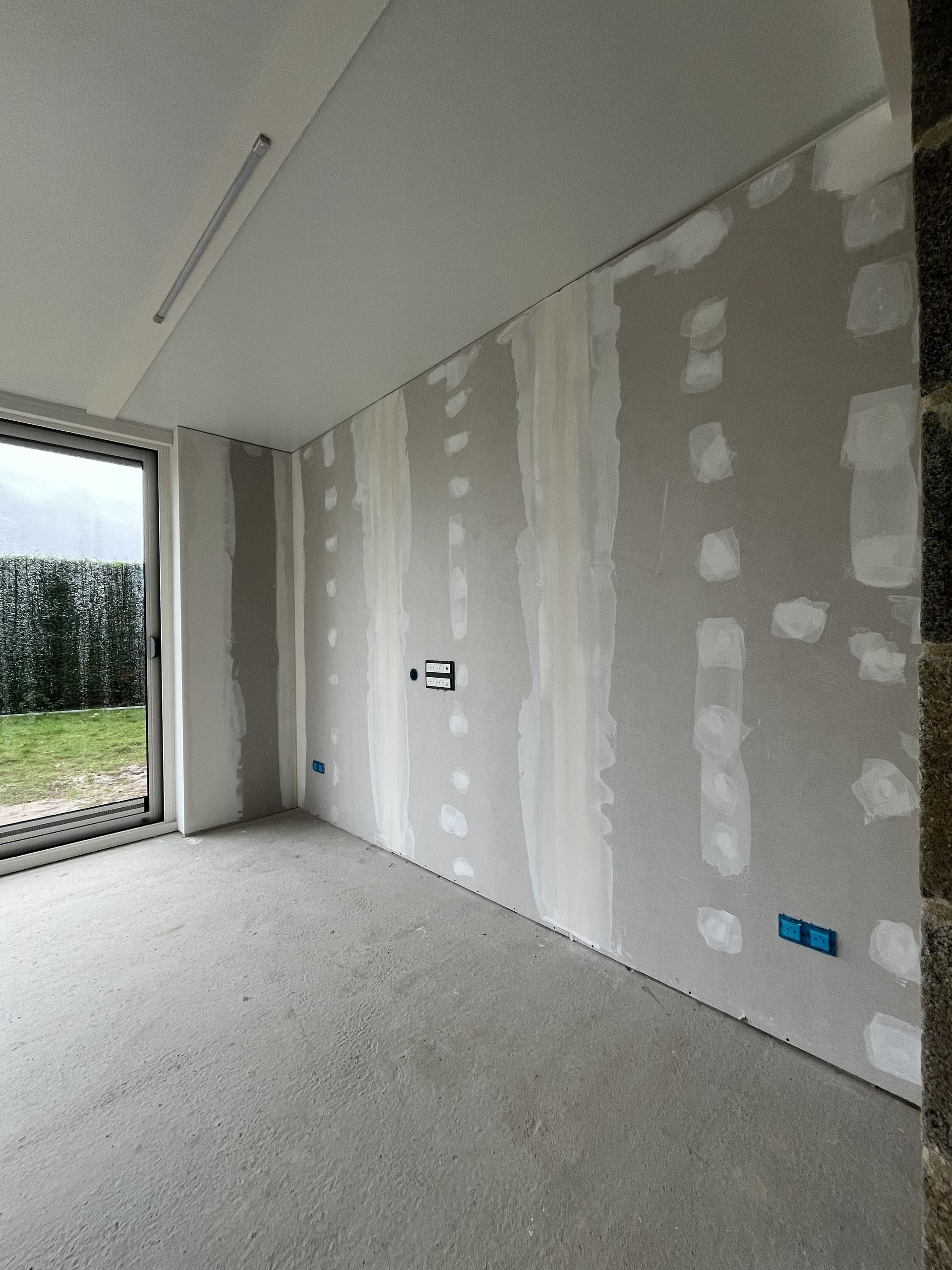 Pièce partiellement aménagée avec des plaques de plâtre réparées, des murs blancs et une grande ouverture fenêtre/porte donnant sur la verdure.