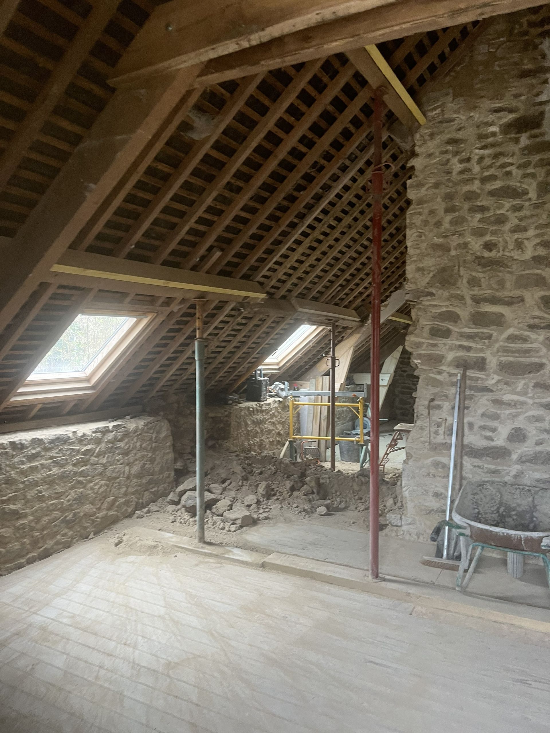 Rénovation d'un grenier en pierre avec poutres apparentes, puits de lumière et échelle adossée au mur.