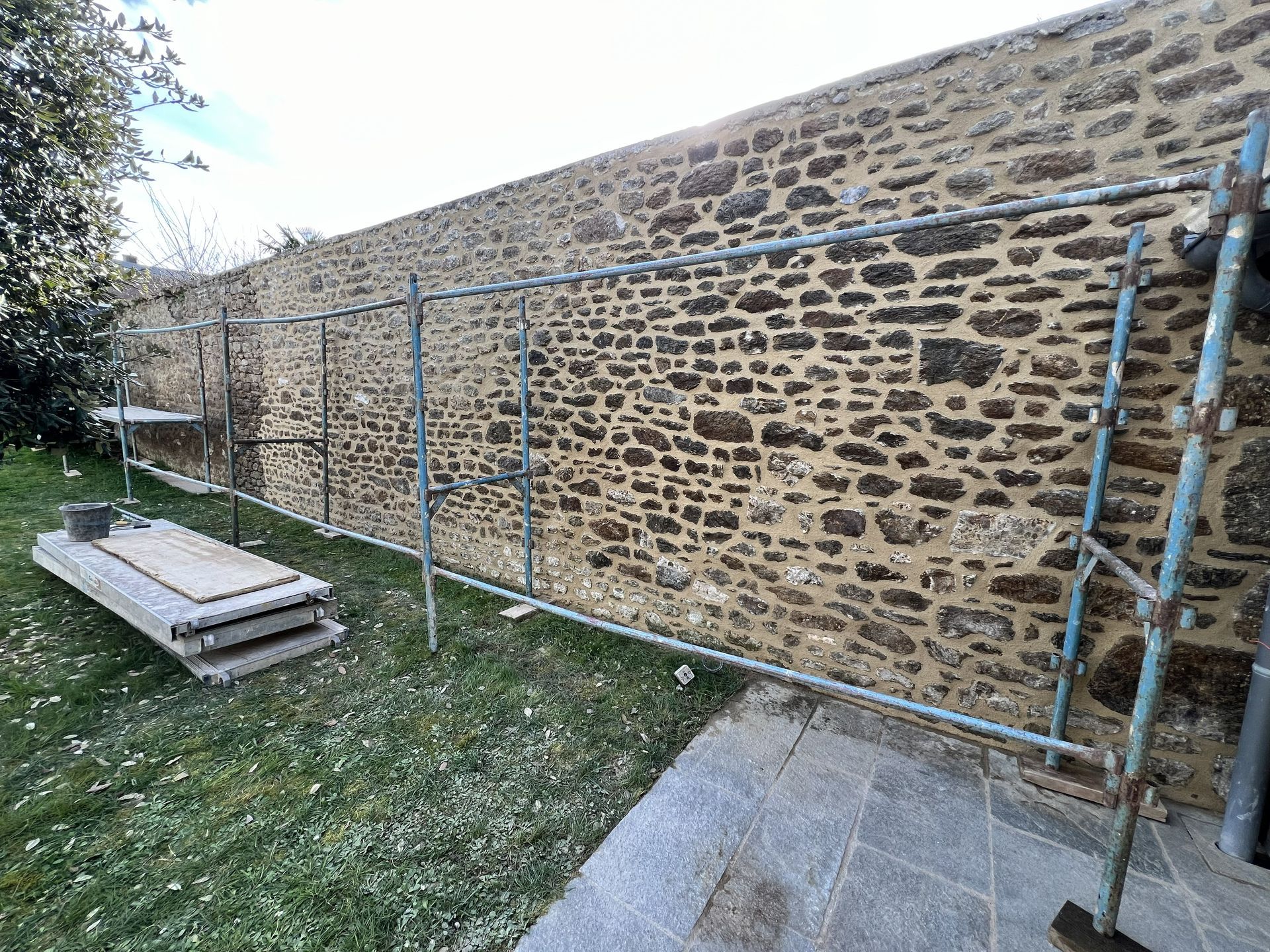 Échafaudage contre un mur de pierre, à côté d'une cour gazonnée et d'un chemin en béton