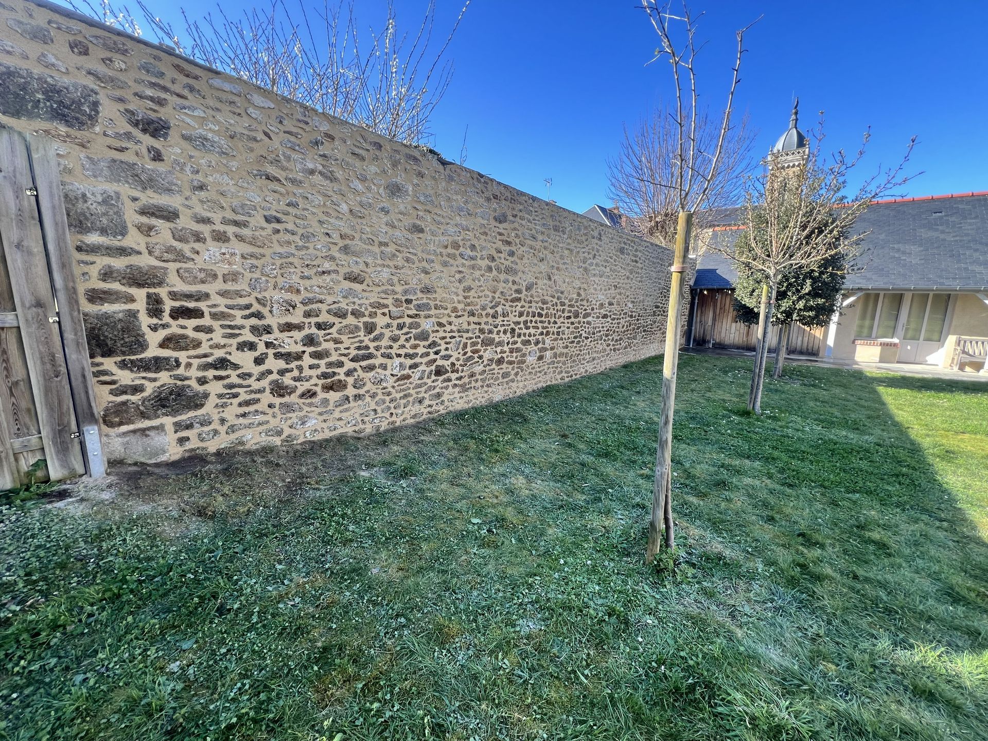 Mur de pierre bordant une cour gazonnée avec de jeunes arbres sous un ciel bleu limpide