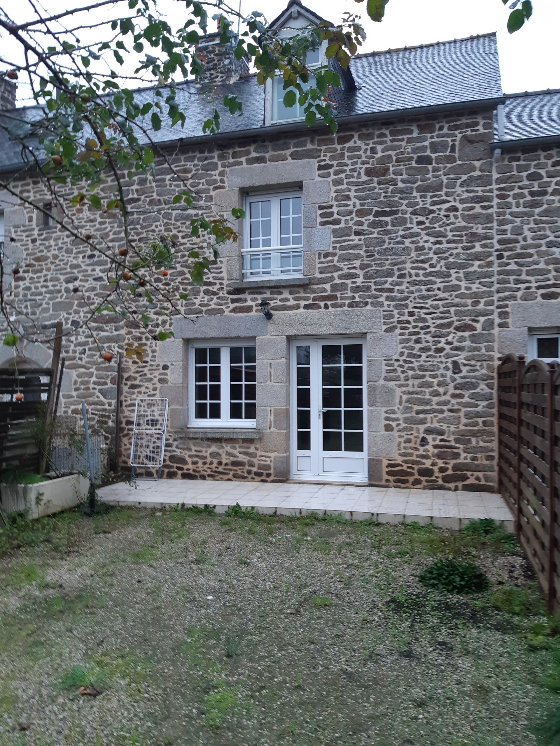 Maison en pierre avec porte et fenêtre blanches, petite cour gravillonnée et branche d'arbre devant.