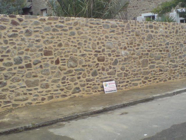 Mur en pierre bordant une rue pavée, avec un petit panneau incliné à sa base.