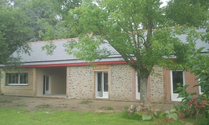 Maison en pierre de plain-pied avec un toit rouge, des portes blanches et de grands arbres dans un jardin gazonné.
