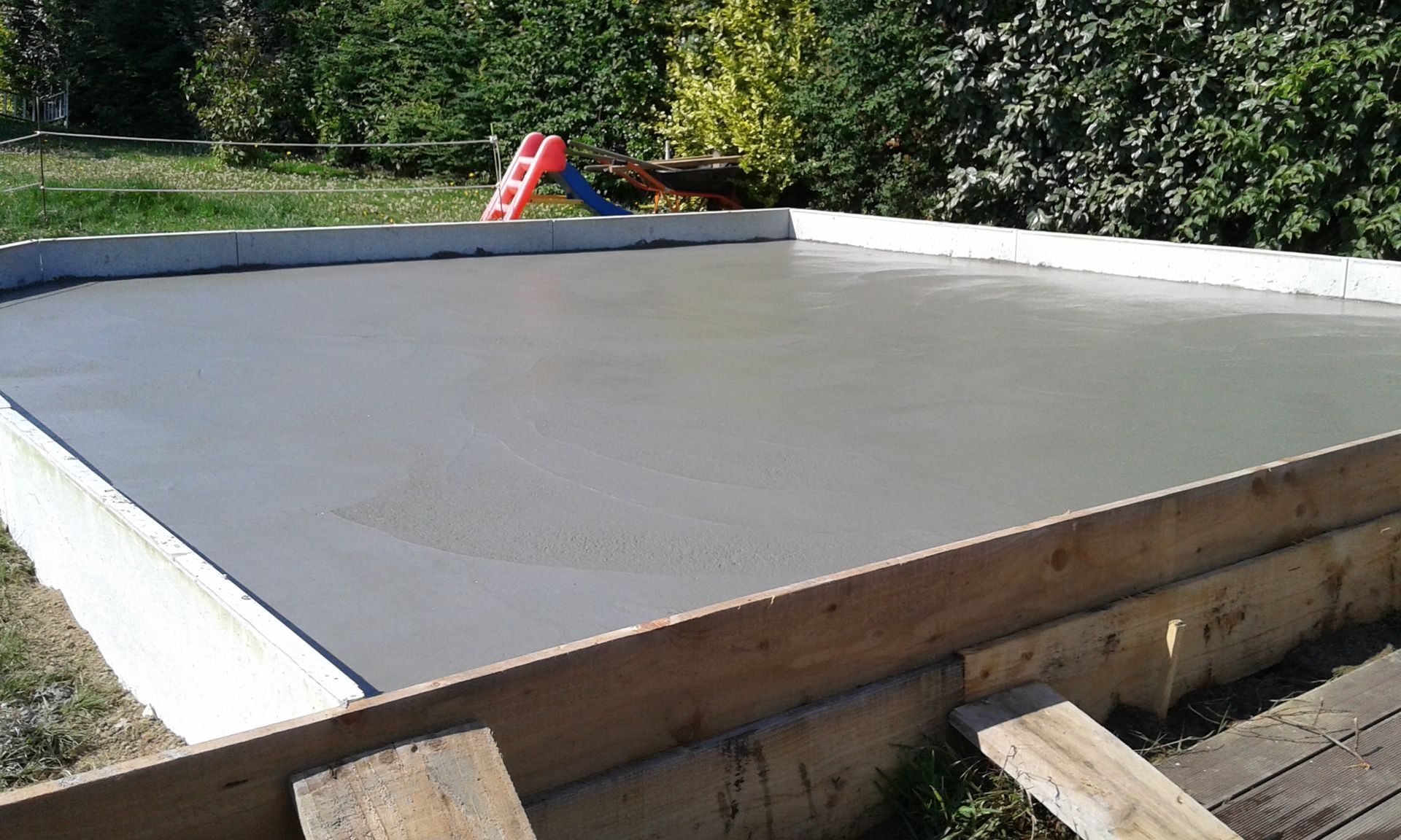 Dalle de béton fraîchement coulée avec coffrage en bois à l'extérieur, entourée de verdure.