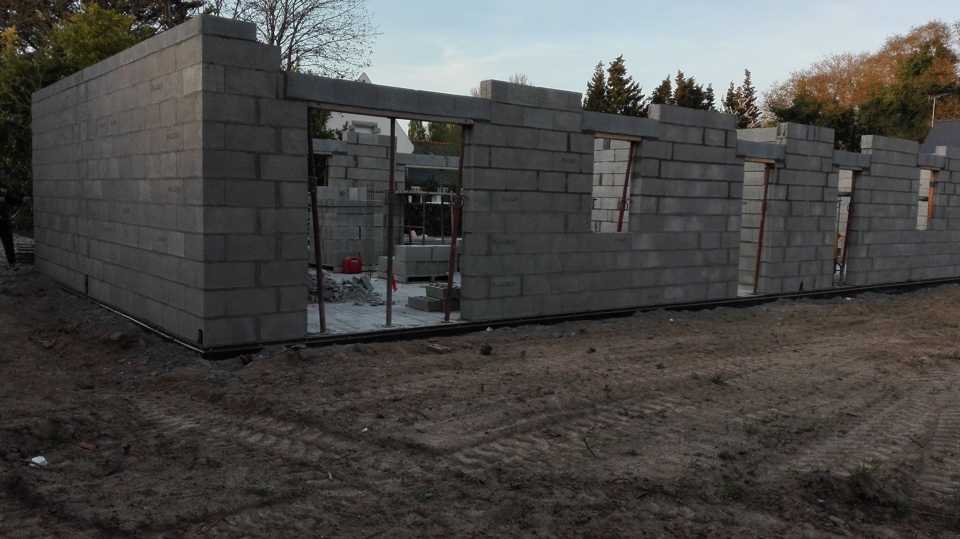 Maison en parpaings partiellement construite, avec des ouvertures de fenêtres inachevées, sur un terrain en terre battue au crépuscule
