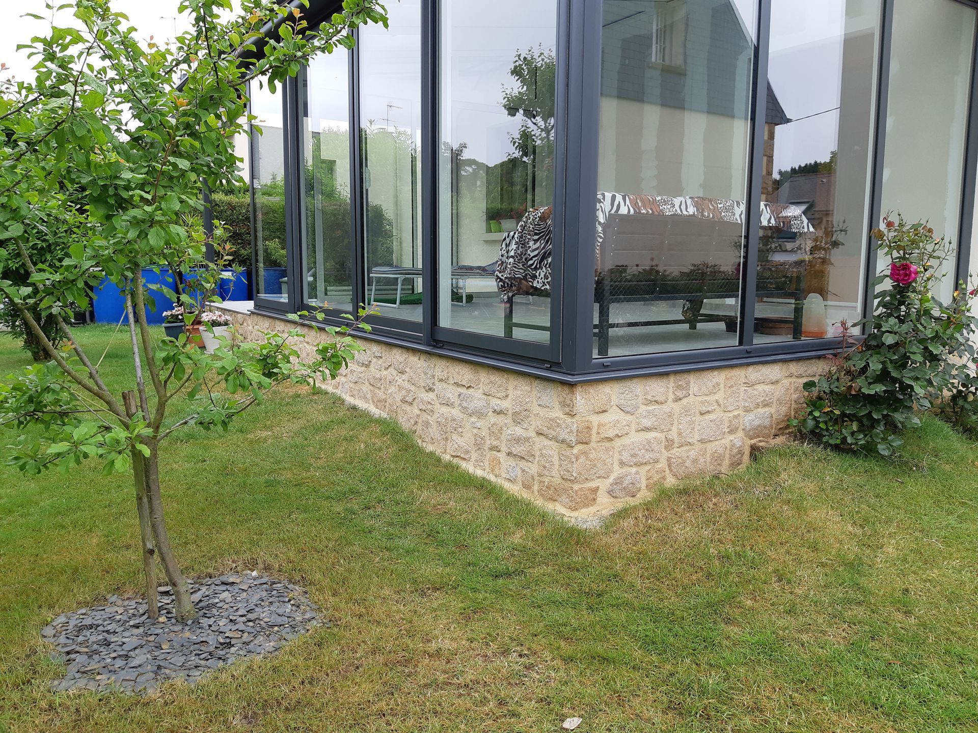 Bâtiment d'angle moderne en verre avec soubassement en pierre, pelouse, jeune arbre et arbustes devant.