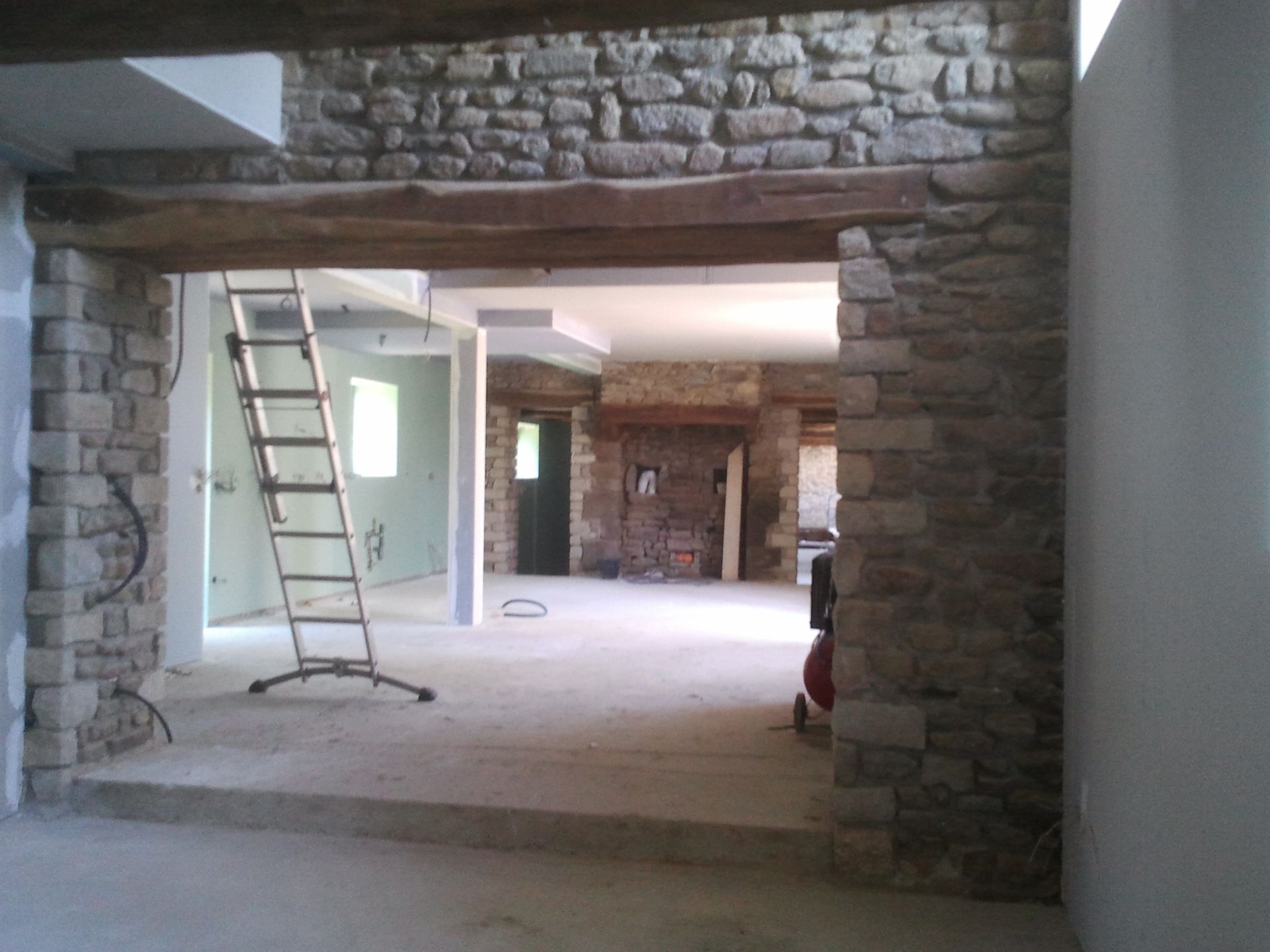 Intérieur aux murs de pierre en cours de rénovation avec une échelle, des poutres apparentes et une porte ouverte