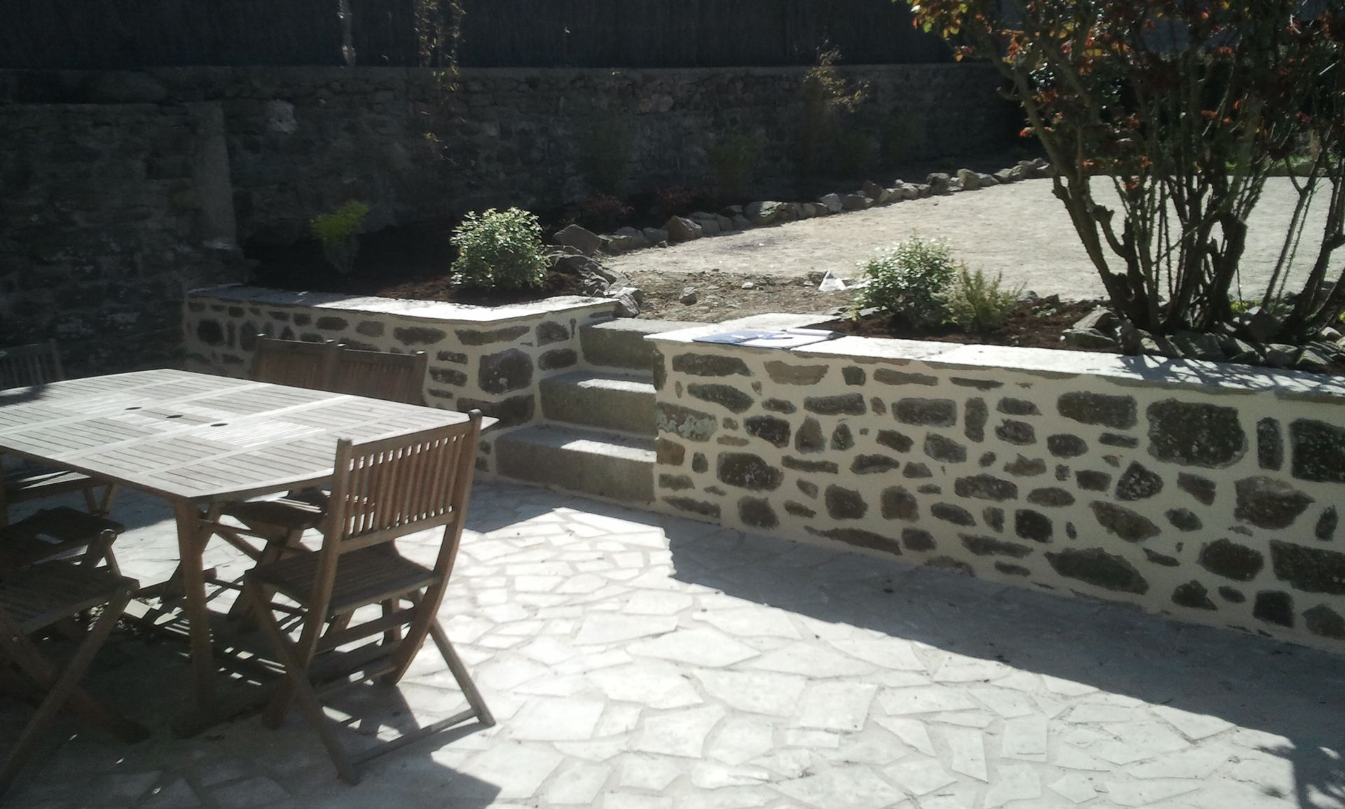 Terrasse ensoleillée avec table et chaises en bois, à côté de jardinières en pierre et d'un muret.