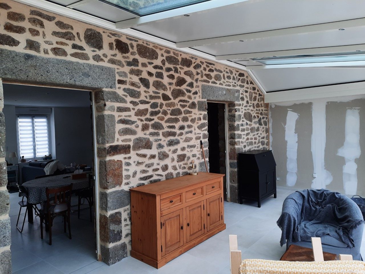 Pièce aux murs de pierre avec portes ouvertes, armoire en bois, poêle noir et chaise recouverte d'une couverture bleue.