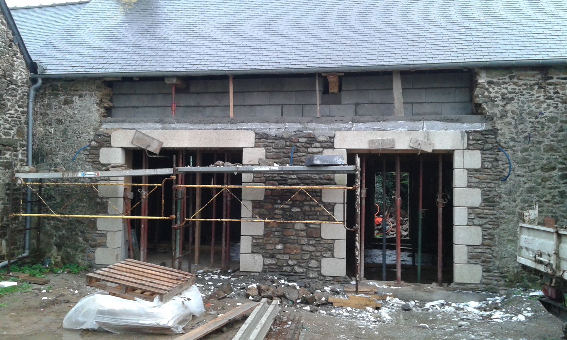 Façade en pierre d'un bâtiment en rénovation, avec échafaudages, ouvertures de portes apparentes et débris de construction au sol.