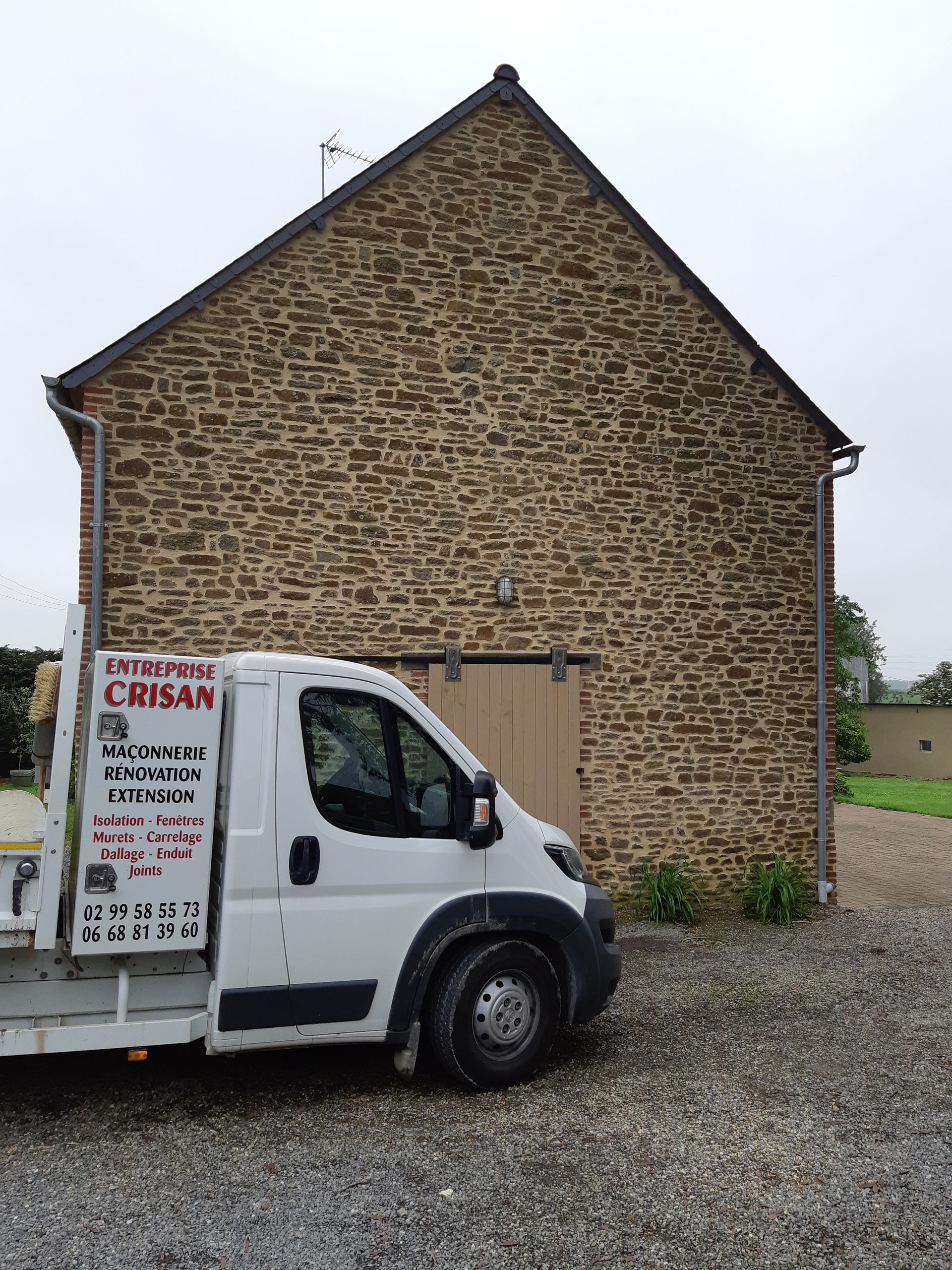 Un camion de livraison blanc est garé devant un bâtiment en pierre au toit pentu et à la cour gravillonnée.