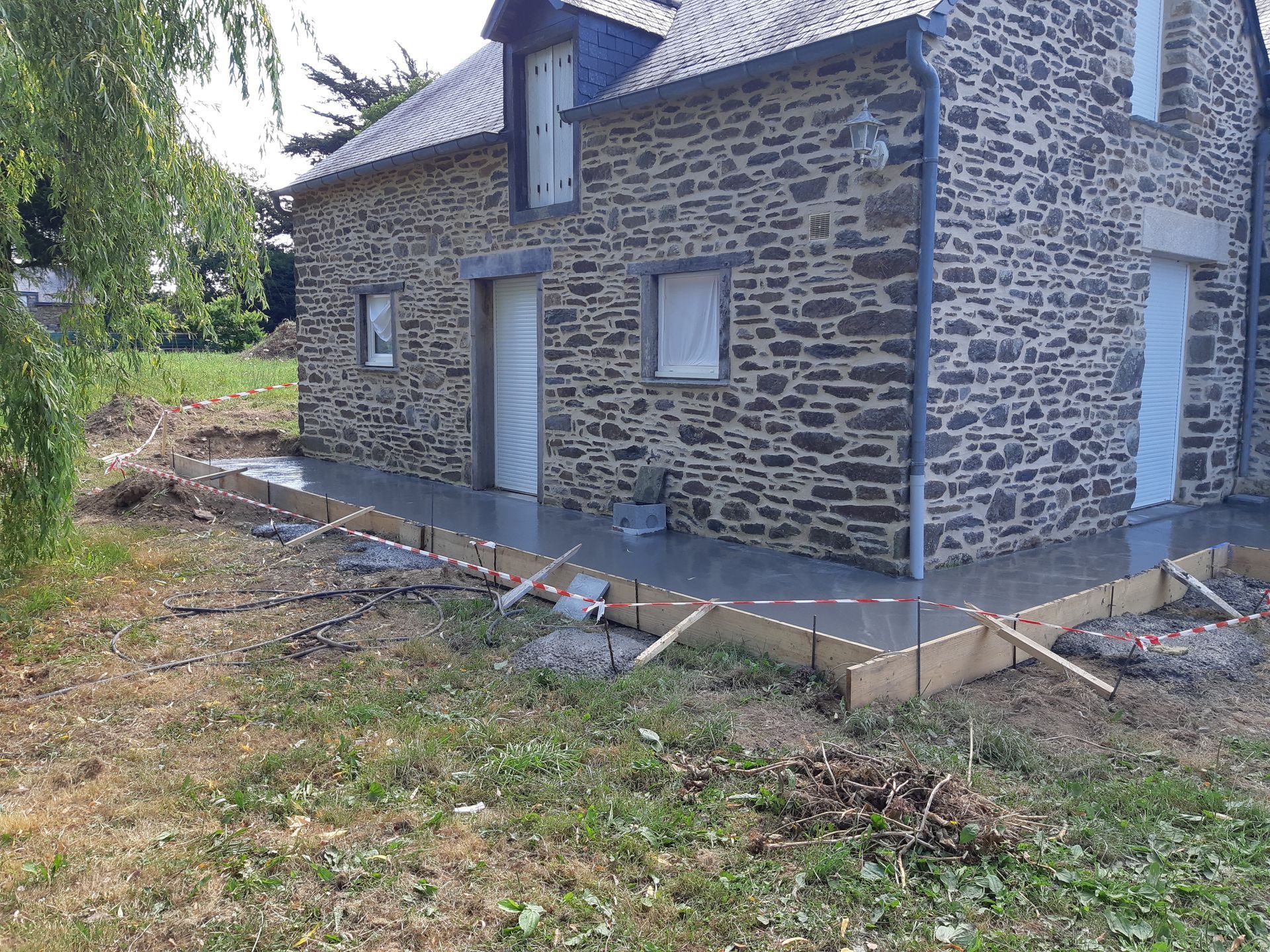 Maison en pierre avec des boiseries bleues et un porche en béton inachevé dans une cour gazonnée