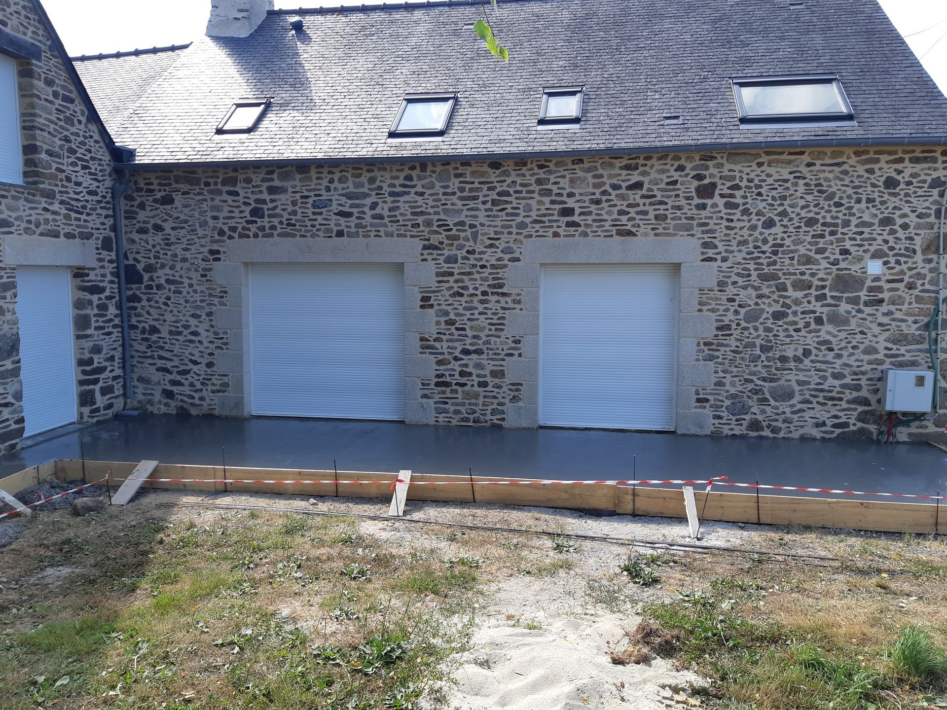 Maison en pierre avec deux portes de garage blanches et une terrasse pavée à l'avant.