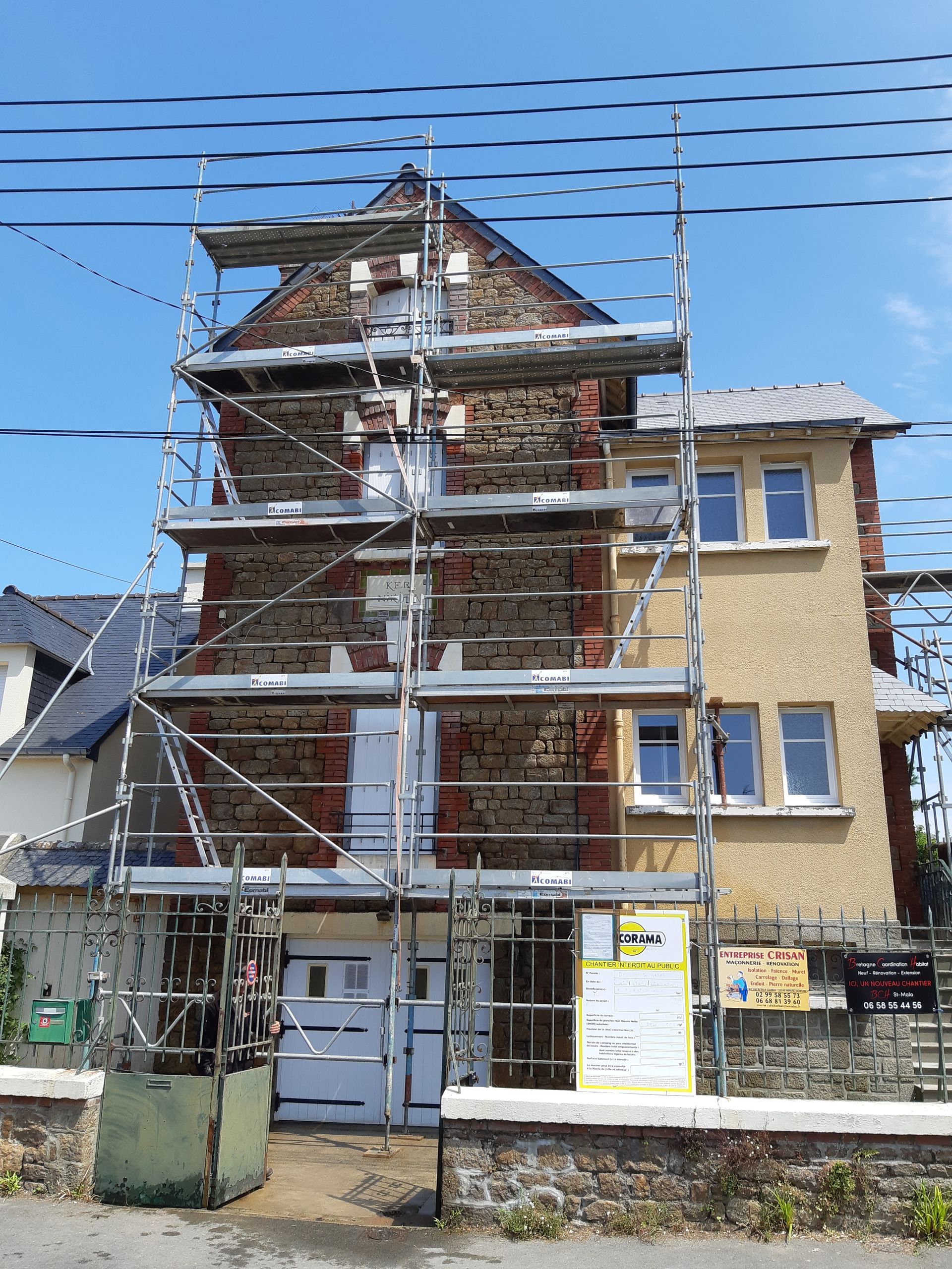 Maison en travaux sous échafaudages, dans une rue ensoleillée, avec une porte blanche et un ciel bleu.