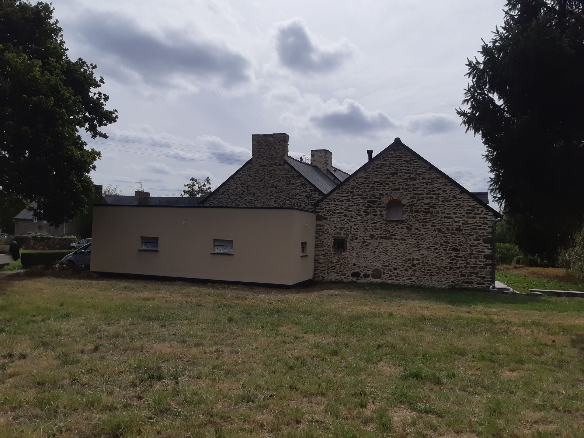 Maison en pierre avec une extension beige dans un champ herbeux sous un ciel nuageux