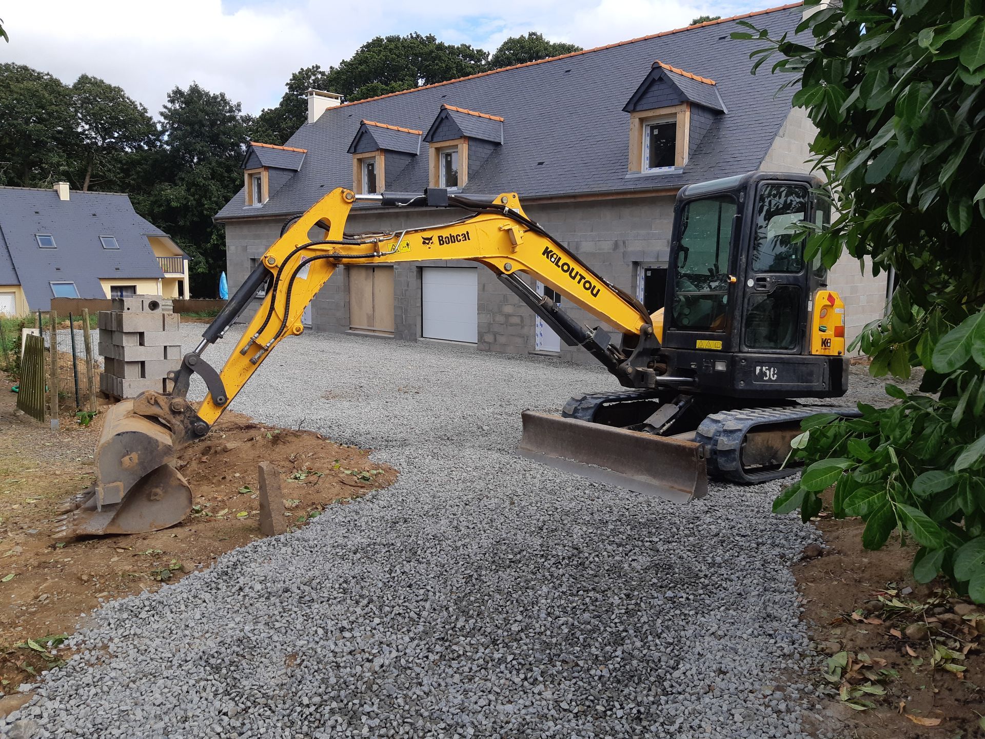 Une pelleteuse répand du gravier sur une allée devant une maison au toit d'ardoise.