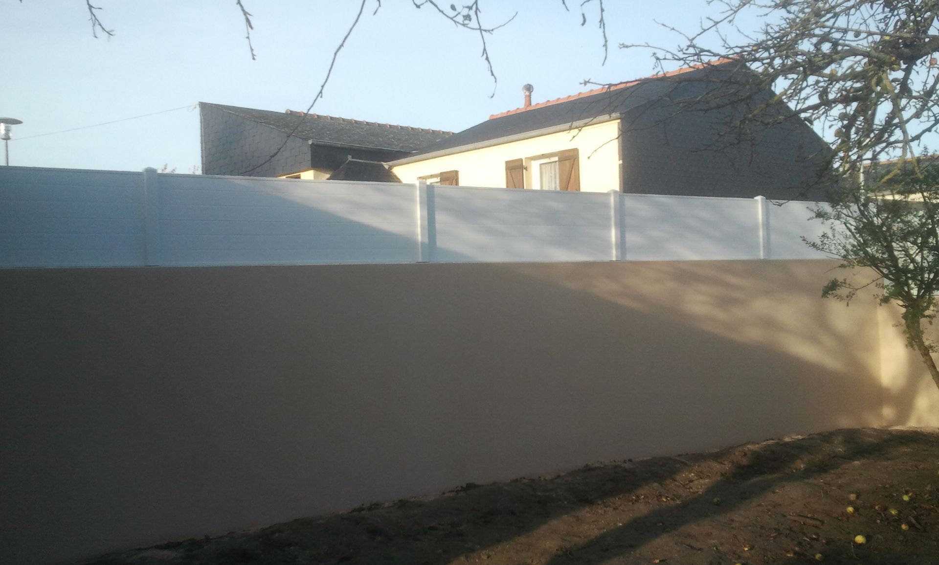 Maison ensoleillée derrière une haute clôture beige et blanche, avec des branches d'arbres dénudées au-dessus