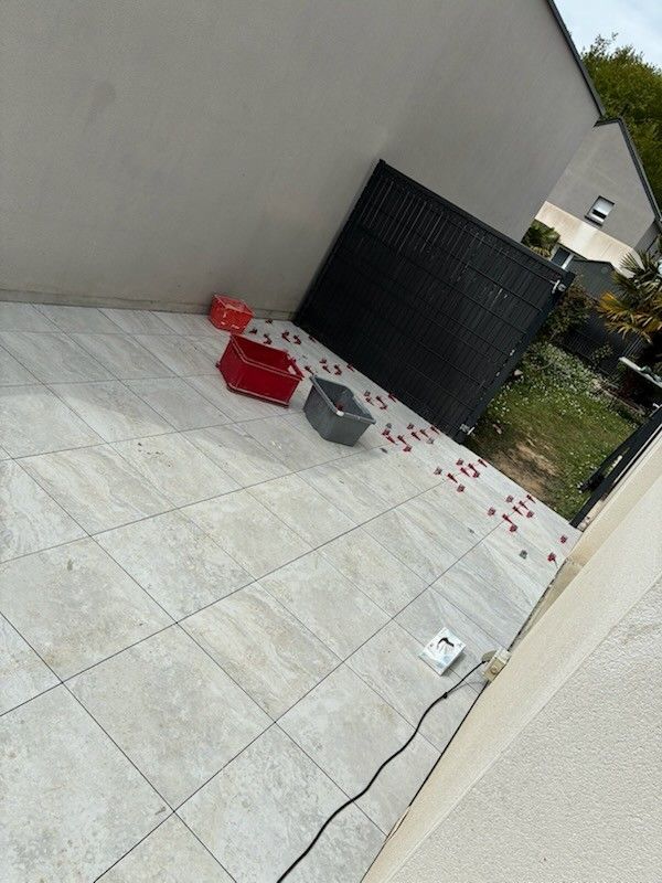 Terrasse avec une glacière rouge renversée et un seau gris sur un sol carrelé, à côté d'un mur et d'un portail noir.