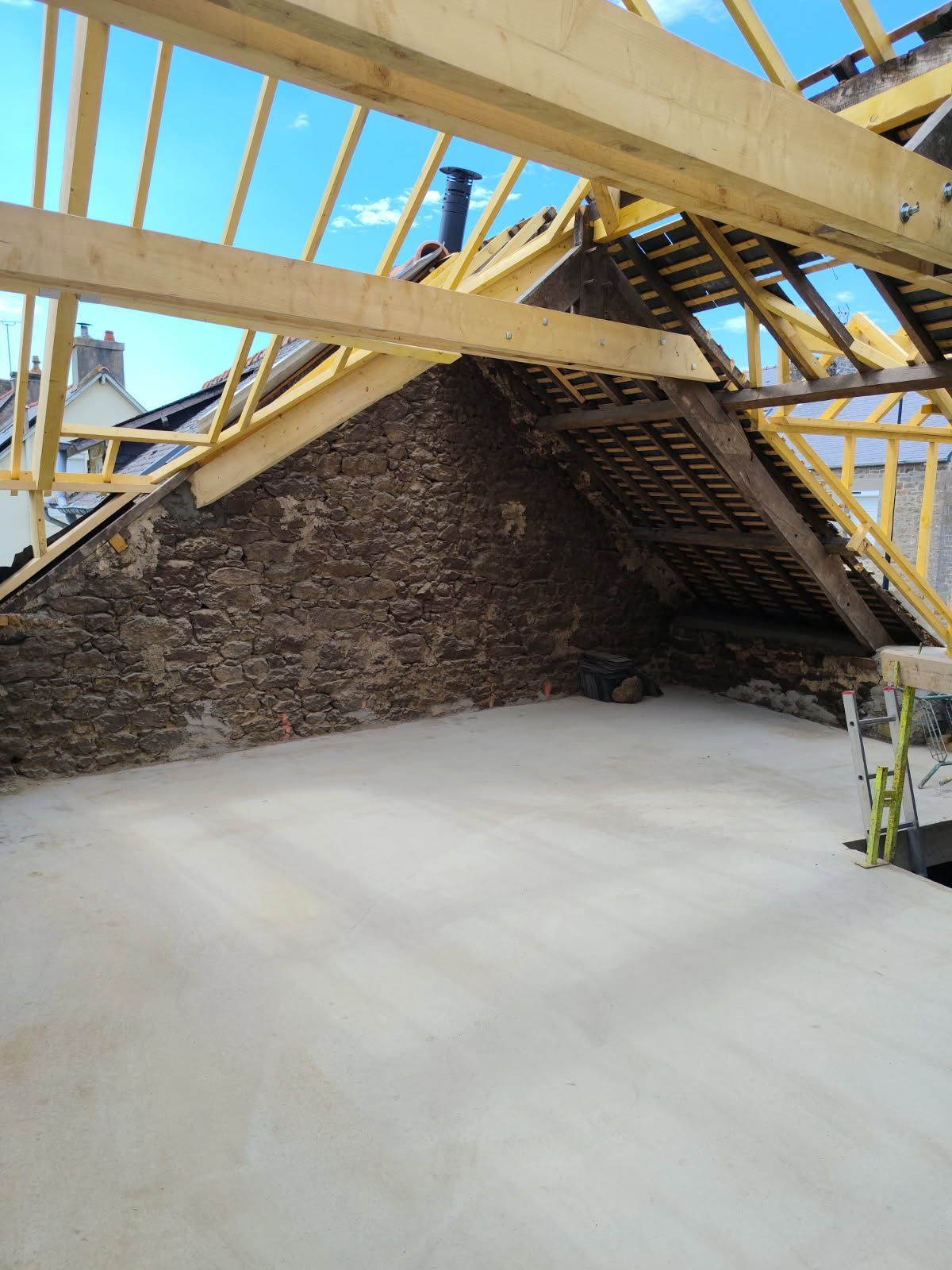 Structure en construction avec charpente apparente en bois et mur pignon en pierre.
