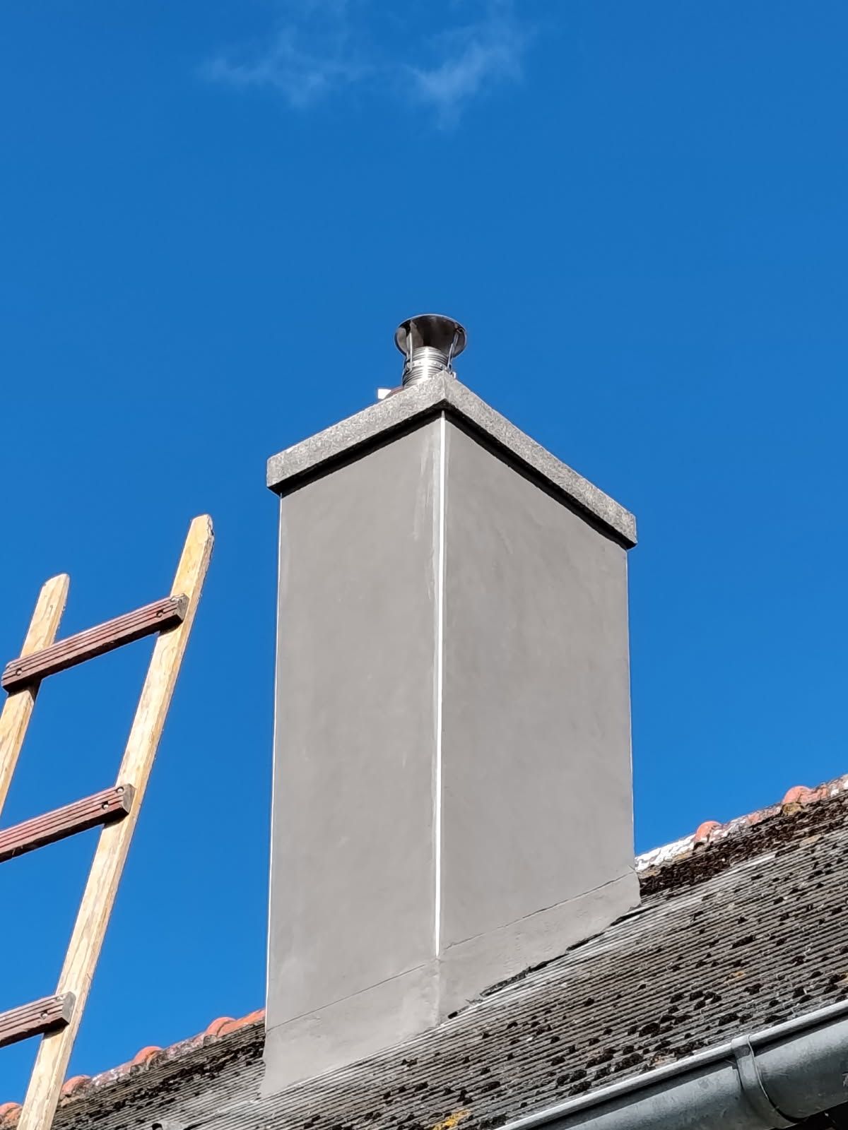 Cheminée grise sur un toit de tuiles, à côté d'une échelle en bois, se détachant sur un ciel bleu clair.
