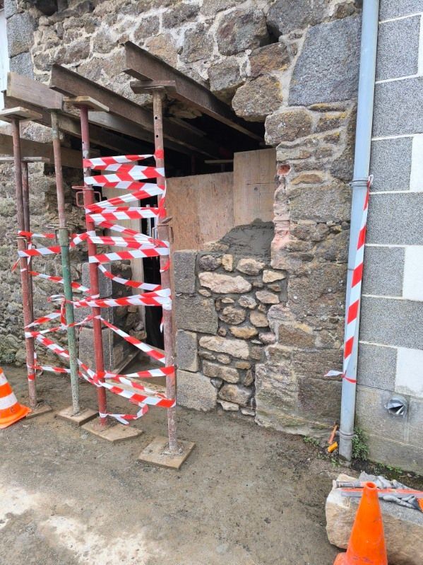 L'entrée d'un bâtiment en pierre avec échafaudages est bloquée par du ruban de signalisation rouge et blanc et des cônes orange.