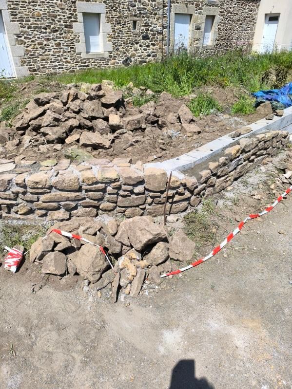 Un mur de pierre effondré jouxte un bâtiment en pierre, avec des décombres et du ruban de signalisation rouge en bord de route.