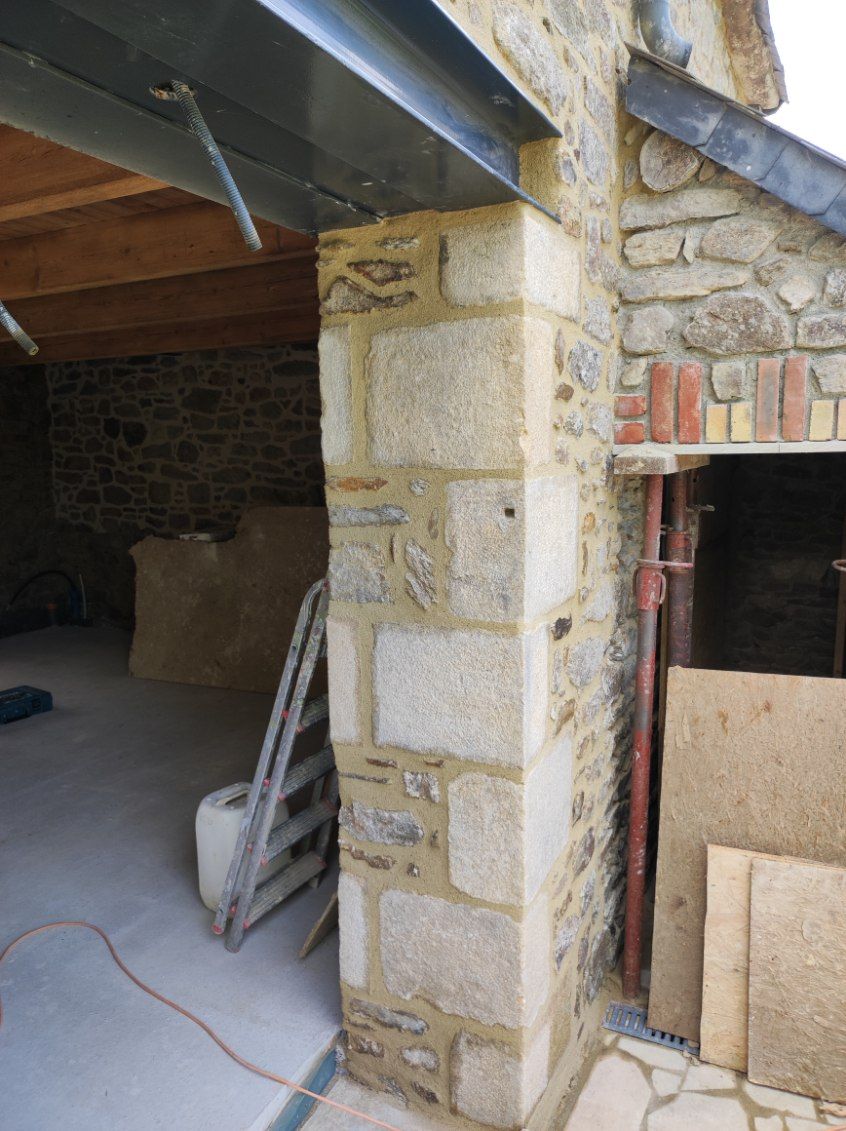 Pièce aux murs de pierre en cours de rénovation, avec poutres apparentes, échelle et porte ouverte recouverte de contreplaqué.