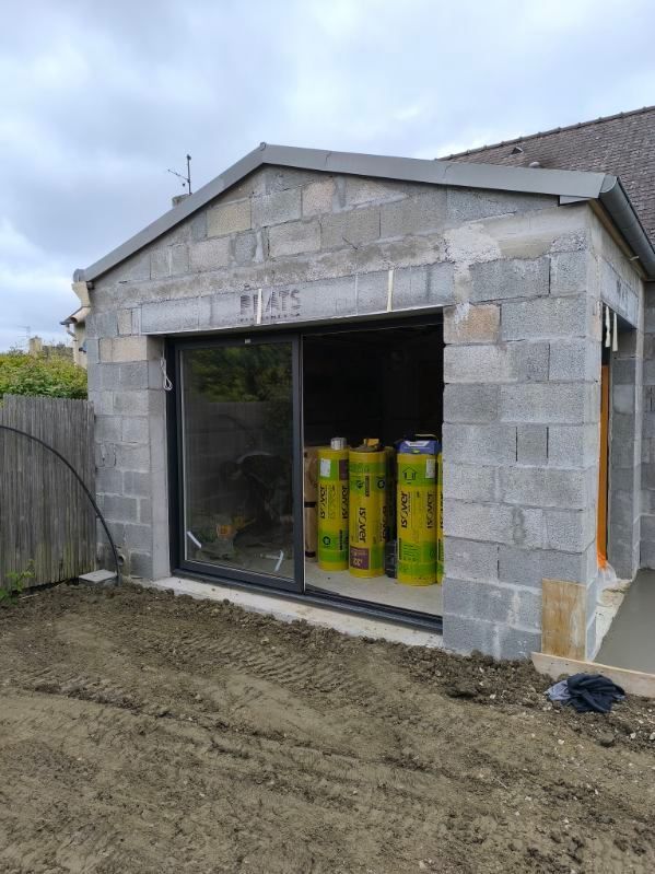 Petit garage en béton brut, avec façade ouverte et matériaux jaune-vert empilés à l'intérieur.