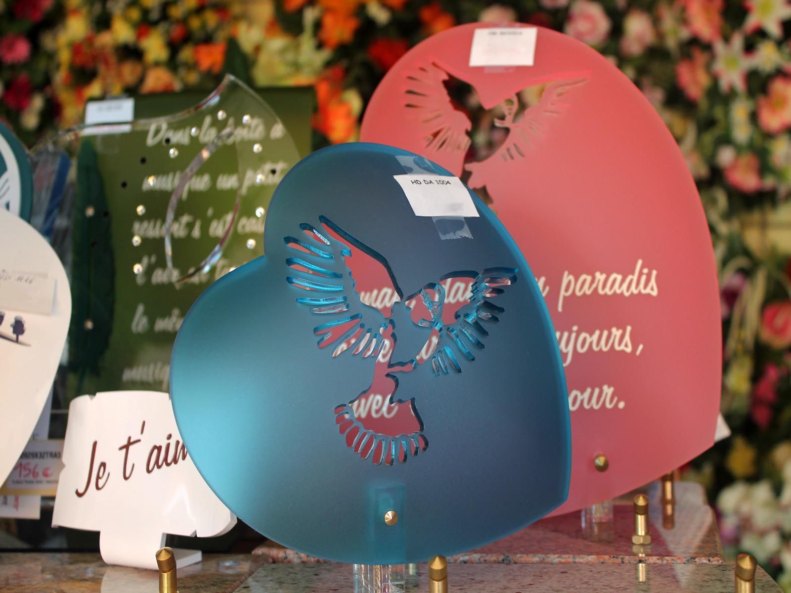 Plaques commémoratives en forme de cœur de différentes couleurs : bleu, vert et rose, avec des motifs gravés et un texte en français.