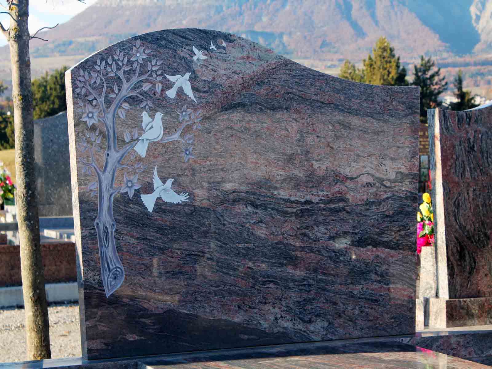 Pierre tombale ornée d'un arbre et de colombes gravés, dans un cimetière avec des montagnes en arrière-plan.