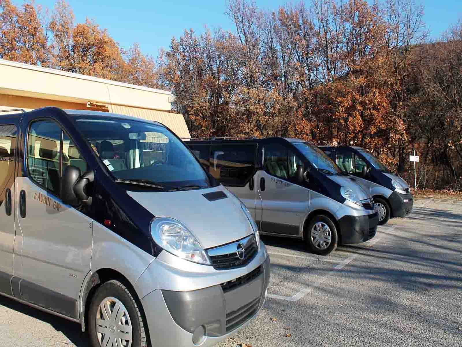Trois minivans argentés garés sur un parking, journée ensoleillée, arbres dénudés en arrière-plan.