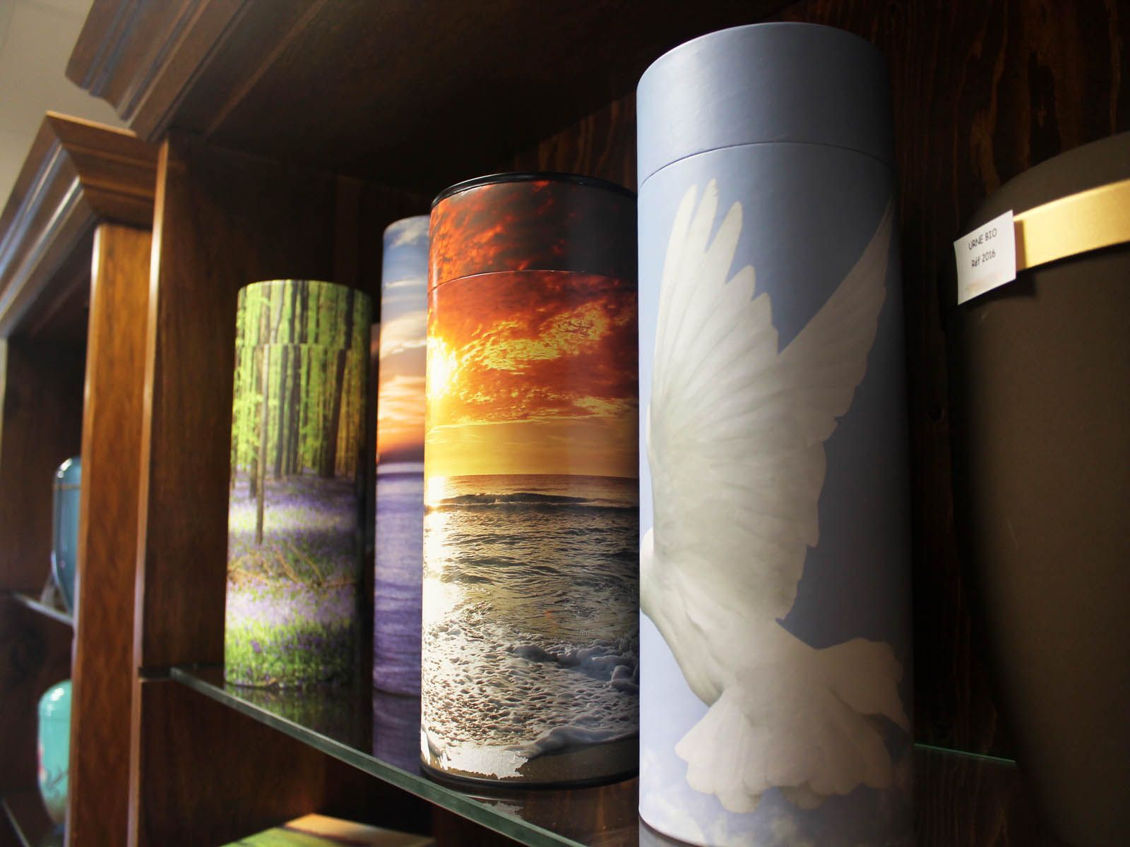 Rangée d'urnes cylindriques ornées de motifs sur le thème de la nature, posées sur une étagère en bois : forêt, coucher de soleil, colombe.