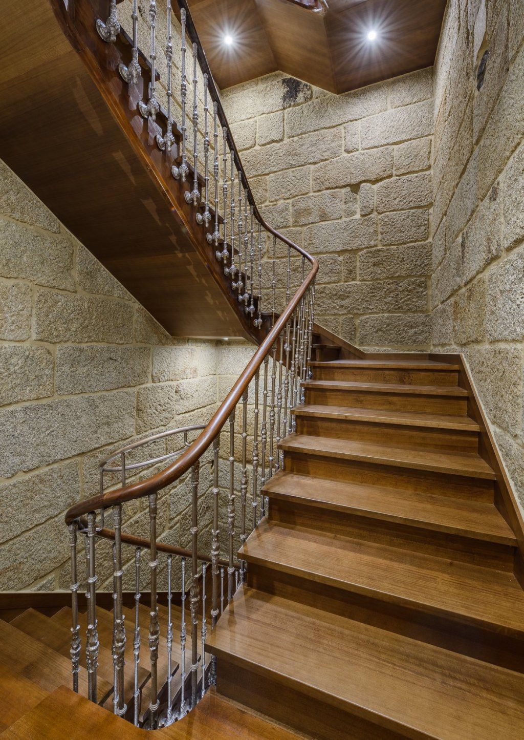 Una escalera de madera con barandilla de metal y una pared de piedra.