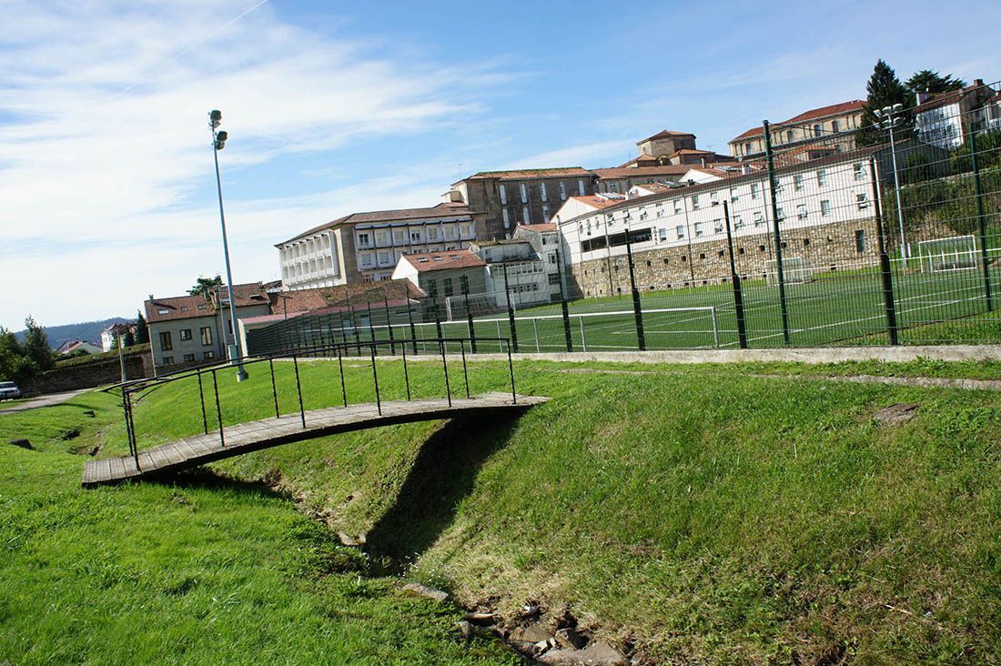Un pequeño puente de madera sobre un arroyo en un parque con edificios al fondo.
