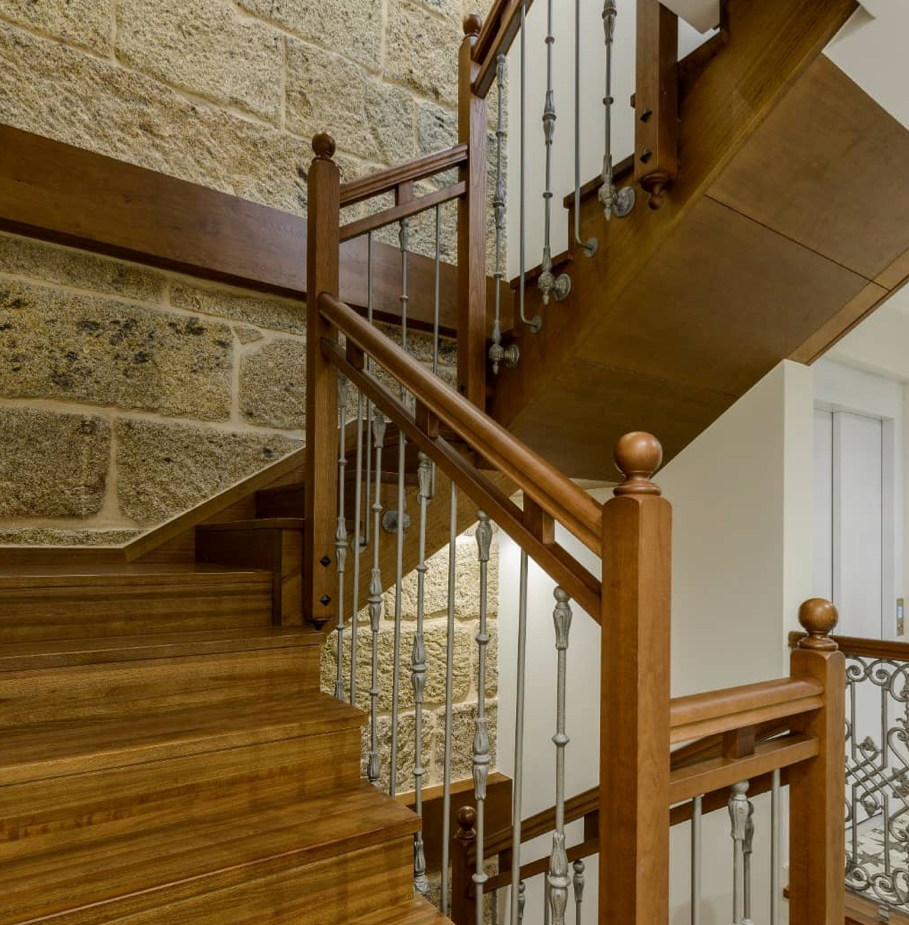 Una escalera de madera con barandilla de metal frente a un muro de piedra.