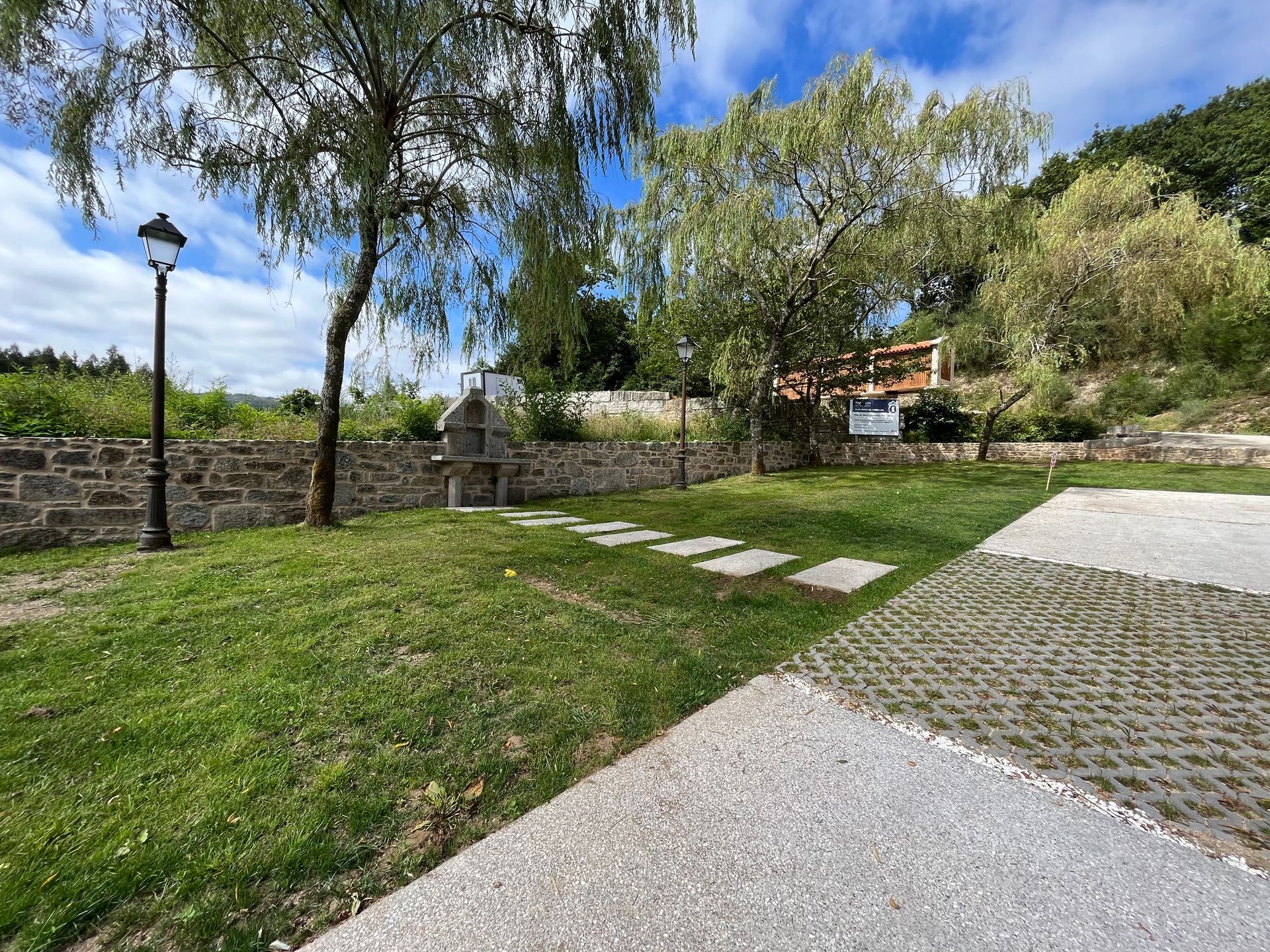 Una pasarela de hormigón que conduce a una zona verde con árboles y un muro de piedra.