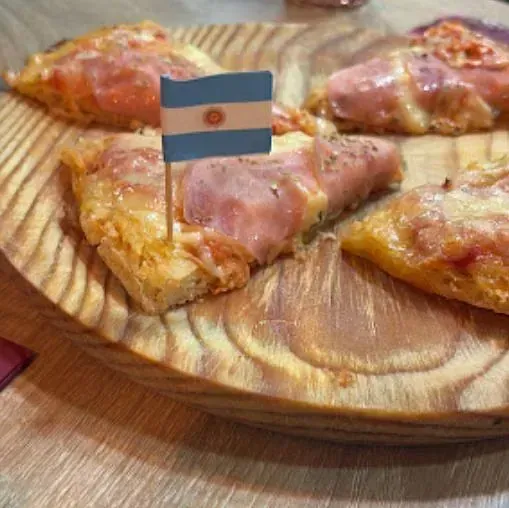 Porciones de pizza sobre una tabla de madera, con un palillo de la bandera argentina. La pizza lleva jamón y queso.