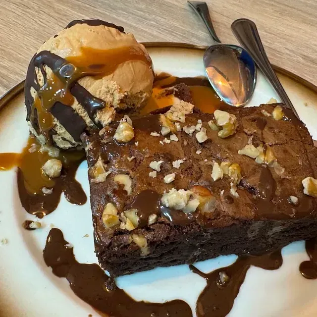 Brownie cubierto con nueces y salsa de chocolate, servido con una bola de helado de caramelo y una cuchara en un plato.
