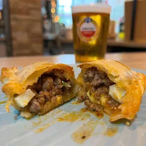 Una empanada cortada para revelar un relleno de carne y huevo en un plato azul, con una cerveza de fondo.