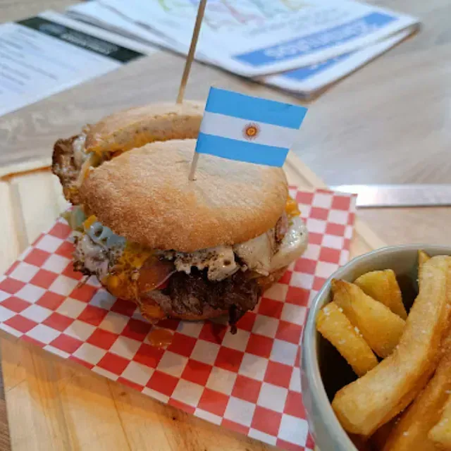 Hamburguesa con palillo de bandera argentina, servida con papas fritas sobre tabla de madera y papel cuadriculado.